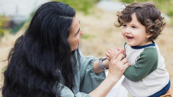 
		Mutter hockt vor ihrem kleinen Kind und greif seine Arme, Kind schaut fröhlich zur Seite
	